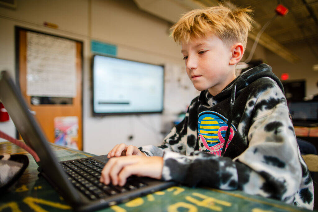 A young boy wearing a black and white tie-dye hoodie uses a laptop at a classroom table. The classroom is bright, with a blurred whiteboard and door in the background.