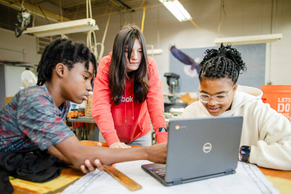 Three students collaborate around a laptop in a classroom. One points at the screen while another smiles and the third leans in to look, with tools and classroom equipment visible in the background.
