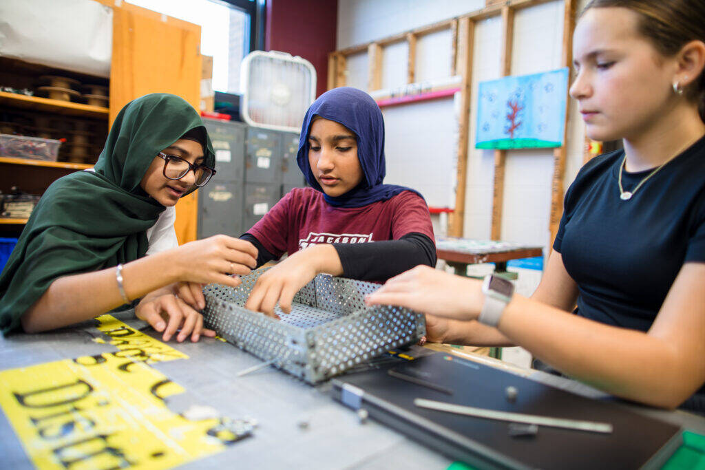 Three students work together assembling a metal structure with bolts and perforated plates in a classroom, focusing intently on their project at a table covered with tools and materials.